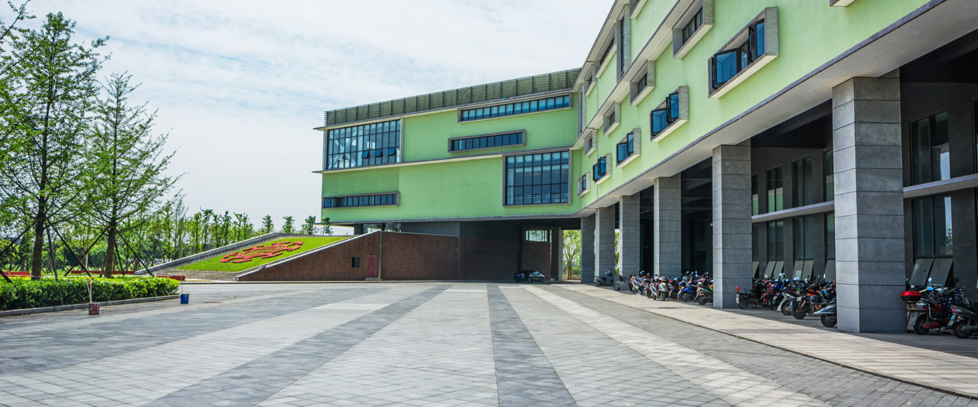 Gedung sekolah bertingkat berdinding hijau muda dengan halaman berpaving luas, taman rumput, dan deretan sepeda motor terparkir di bawahnya