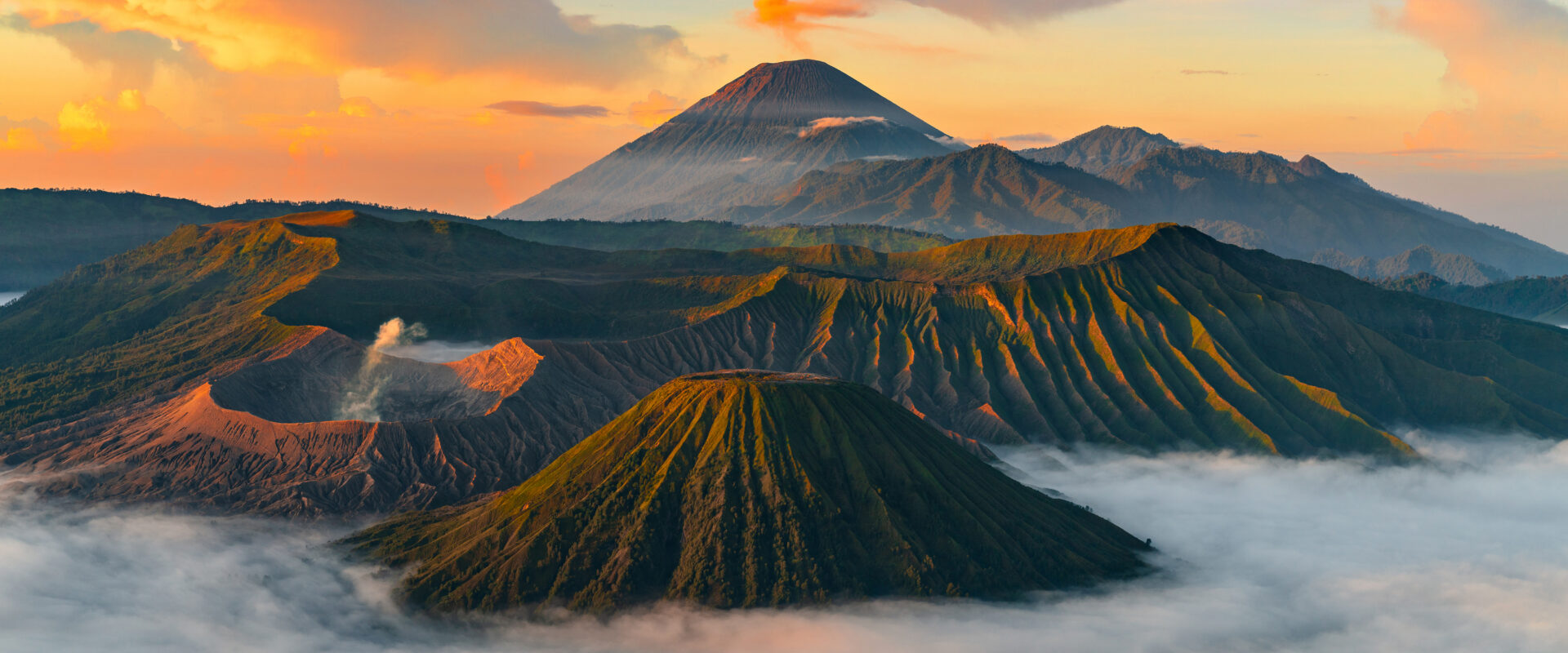 Lanskap pegunungan dengan lautan kabut di lembah dan puncak kerucut menjulang di kejauhan, di bawah langit oranye saat matahari terbit